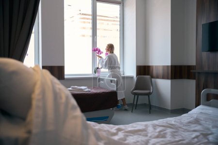 Rückenansicht eines Patienten im weißen Bademantel, der Orchideenduft inhaliert, der auf der Fensterbank der Krankenstation platziert wird