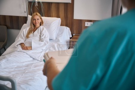 Lächelnde Patientin auf Krankenhausbett sitzend und Krankenschwester mit abgedecktem Tablett in der Hand anschauend