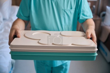 Cropped photo of male nurse holding covered food tray in hands while standing near hospital bed
