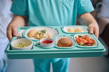 Foto de Foto recortada del asistente de enfermería sosteniendo bandeja de comida en las manos mientras está de pie en la habitación del hospital - Imagen libre de derechos