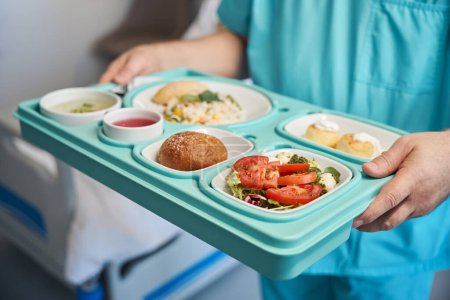 Cropped photo of nursing assistant with plastic food tray in hands standing beside hospital bed