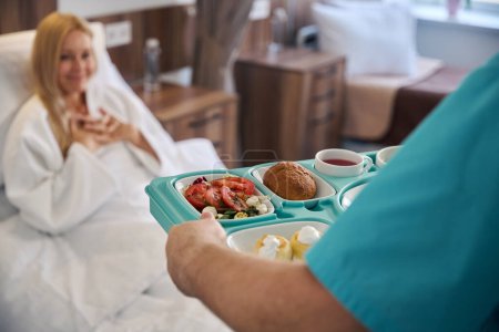 Nursing assistant holding plastic food tray in front of hospital bed with recumbent female patient