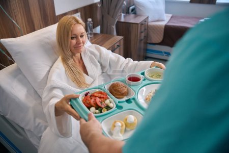 Contented female patient seated in hospital bed taking food tray from hands of nursing assistant