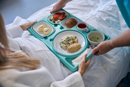 Foto de Foto recortada de manos de enfermero dando bandeja de comida a una paciente hospitalizada sentada en la cama del hospital - Imagen libre de derechos