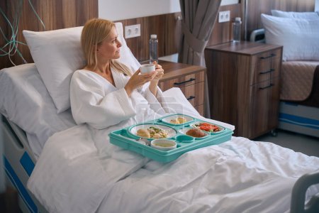 Nachdenkliche Stationärin sitzt im Krankenhausbett mit einem Tablett mit Essen, hält eine Tasse Tee in den Händen und schaut weg