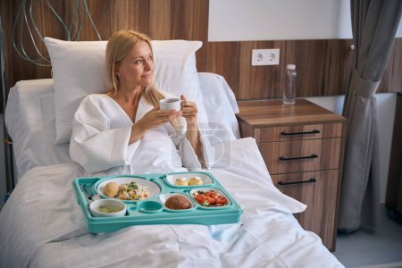 Téléchargez les photos : Patient pensif assis dans un lit d'hôpital avec plateau alimentaire tenant une tasse de boisson à base de plantes dans les mains et regardant de côté - en image libre de droit