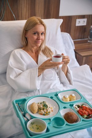 Paciente femenina contenta sentada en la cama del hospital con bandeja de comida sosteniendo taza de bebida herbal en las manos