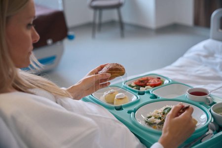 Patientin sitzt im Krankenhausbett und hält Brötchen in der Hand, während sie Salat mit Gabel isst