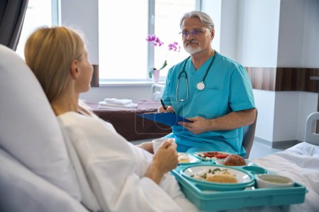 Téléchargez les photos : Médecin traitant sérieux avec presse-papiers et stylo dans les mains regardant un patient hospitalisé assis sur un lit d'hôpital pendant le déjeuner - en image libre de droit
