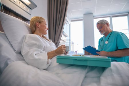 Médico serio tomando notas en el portapapeles con pluma en presencia de una paciente sentada en la cama del hospital con bandeja de comida