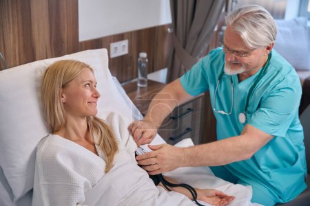 Lächelnde Frau im Krankenhaus liegt im Bett, während Arzt BP-Manschette um ihren Oberarm wickelt