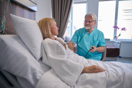 Téléchargez les photos : Patiente couchée dans un lit d'hôpital alors que le médecin mesure sa tension artérielle à l'aide d'un stéthoscope et d'un sphygmomanomètre - en image libre de droit