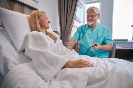 Téléchargez les photos : Patiente hospitalisée allongée dans un lit d'hôpital alors que le clinicien mesure sa tension artérielle avec un sphygmomanomètre et un stéthoscope - en image libre de droit