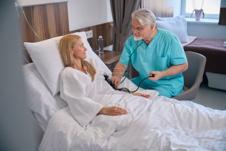Téléchargez les photos : Femme hospitalisée allongée dans un lit d'hôpital tout en prenant soin d'un médecin mesurant sa tension artérielle à l'aide d'un sphygmomanomètre et d'un stéthoscope - en image libre de droit