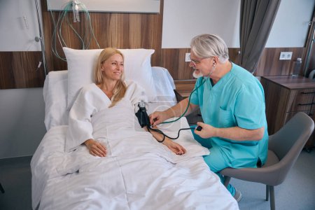 Lächelnde stationäre Patientin liegt im Krankenhausbett, während der behandelnde Arzt während der Stationsrunde ihren Blutdruck misst