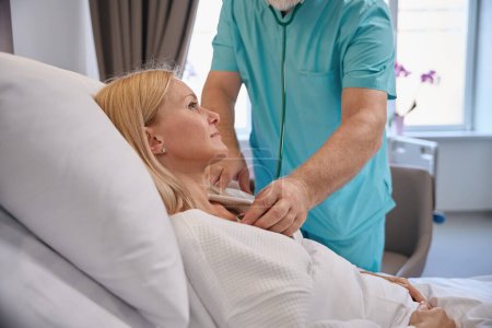 Patientin liegt im Krankenhausbett, während der behandelnde Arzt während der körperlichen Untersuchung ihrem Herzschlag mit Stethoskop zuhört