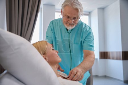 Mujer hospitalizada acostada en la cama del hospital mientras el médico escucha sus latidos del corazón usando estetoscopio durante el examen físico