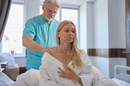 Téléchargez les photos : Femme hospitalisée assise sur le lit d'hôpital pendant que le pneumologue écoute ses bruits pulmonaires avec stéthoscope pendant l'examen physique - en image libre de droit