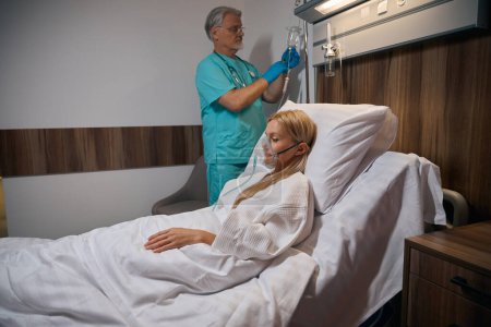 Female patient lying in hospital bed while doctor administering medication with syringe into saline bottle hanging on IV pole hook