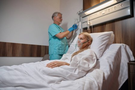 Téléchargez les photos : Femme hospitalisée allongée dans un lit d'hôpital alors que le médecin administre un médicament avec une seringue dans une bouteille saline accrochée au crochet de la perche IV - en image libre de droit