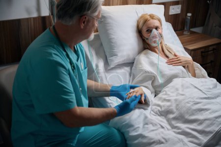 Téléchargez les photos : Médecin traitant tenant la main d'un patient hospitalisé avec masque à oxygène couché dans un lit d'hôpital en salle pendant la perfusion intraveineuse - en image libre de droit