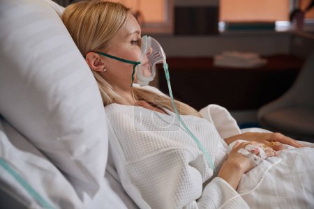 Foto de Paciente en máscara de oxígeno y con catéter intravenoso periférico insertado en vena en la mano acostado en cama de hospital en sala - Imagen libre de derechos