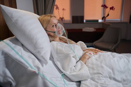Foto de Mujer hospitalizada en máscara de oxígeno y con catéter intravenoso periférico insertado en la vena de su mano acostada en la cama del hospital - Imagen libre de derechos
