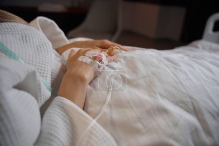Ausgeschnittenes Foto einer kranken Person mit peripheren intravenösen Katheter, der in eine Vene an der Hand eingeführt wird, die im Krankenhausbett liegt