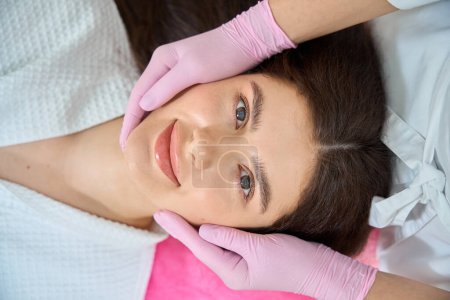 Photo for Top view of young woman lying supine while cosmetologist massaging her cheeks with hands in nitrile gloves - Royalty Free Image