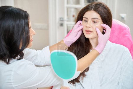 Woman seated in chair looking at herself in hand mirror while dermatologist in nitrile gloves palpating her cheeks
