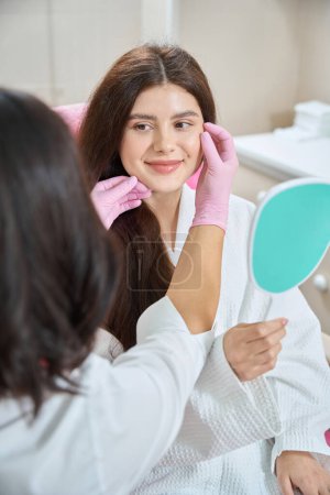 Lächelnde junge Frau sitzt mit Spiegel in der Hand im Stuhl und schaut den Arzt an, der ihre Gesichtshaut während der Konsultation untersucht