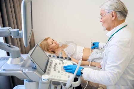 Téléchargez les photos : Médecin masculin déplaçant un transducteur d'ultrasons sur l'abdomen du patient tout en la regardant, effectuant un examen médical et des diagnostics - en image libre de droit