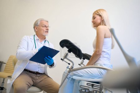 Téléchargez les photos : Femme assise dans une chaise de gynécologie, homme médecin en gants bleus assis à proximité et tenant une liste de contrôle - en image libre de droit