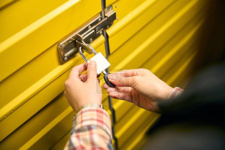 Primer plano de las manos femeninas insertando la llave en el orificio de la cerradura de metal que cuelga en el pestillo de la puerta del contenedor