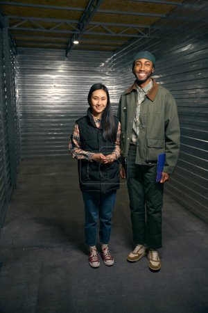 Full-length portrait of cheerful storehouse employee with clipboard and smiling tenant with door lock in hand standing in cargo container