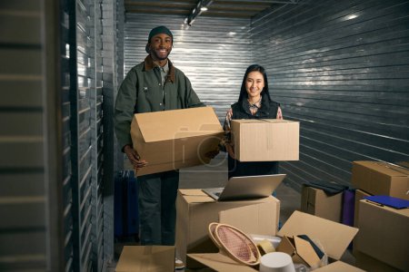 Téléchargez les photos : Travailleurs d'entrepôt souriants avec des boîtes en carton dans les mains debout à l'intérieur du conteneur de stockage et regardant vers l'avenir - en image libre de droit