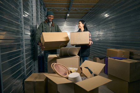 Trabajador de almacén alegre y su compañera de trabajo sonriente sosteniendo cajas de cartón en las manos mientras está de pie en un contenedor de carga