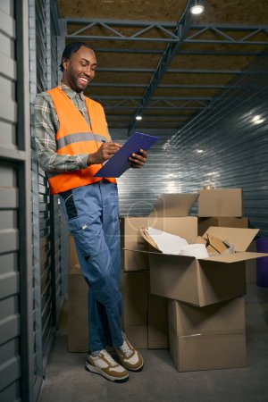 Full-size portrait of pleased worker writing with pen on clipboard while leaning against wall in cargo container