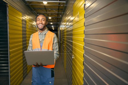Smiling employee with laptop in hands standing in aisle among closed cargo containers