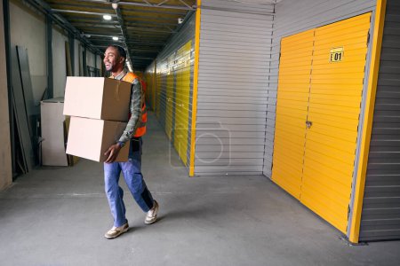 Full-size portrait of merry freight handler carrying pair of cardboard boxes in storage area