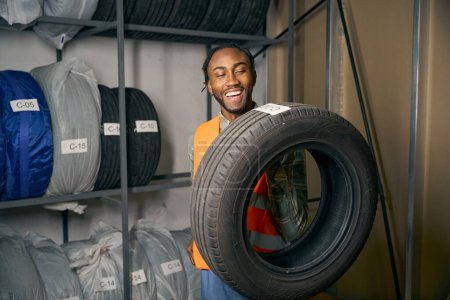 Joyous guy in uniform holding rubber car tire with hand while standing in front of steel storage rack