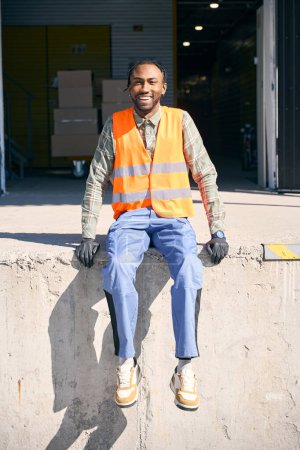 Cheerful young freight handler sitting on concrete loading dock located in front of warehouse facility and looking ahead