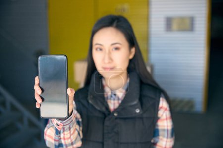 Téléchargez les photos : Portrait d'une jeune ouvrière sérieuse posant avec un téléphone portable dans sa main contre un conteneur de rangement - en image libre de droit