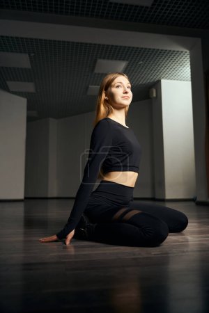 Photo for Slim choreographer dressed in black attire sitting with knees bent on floor during floor dance session - Royalty Free Image