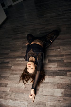 Flexible junge Frau in High Heels und schwarzer Kleidung, die rückwärts gleitet, während sie auf dem Boden liegt