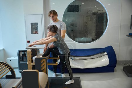 Rehabilitation therapist works with female patient on a special simulator, people in comfortable training clothes