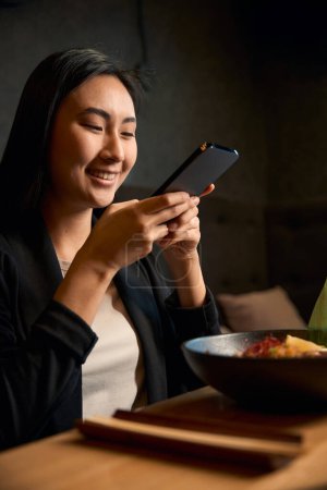 Mujer alegre satisfecho con su deliciosa comida sabrosa y tomar la foto del tazón con ramen de mariscos en el teléfono inteligente para compartir con amigos