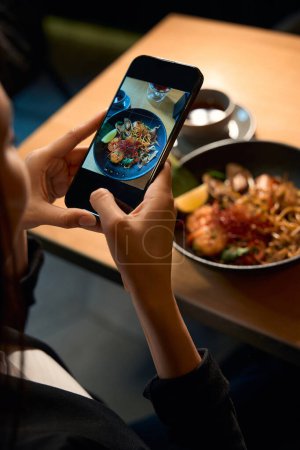 Blick von oben auf die weibliche Smartphone-Kamera, Besucherin eines asiatischen Restaurants beim Fotografieren perfekt servierter und köstlicher Meeresfrüchte-Ramen, die in sozialen Netzwerken geteilt werden