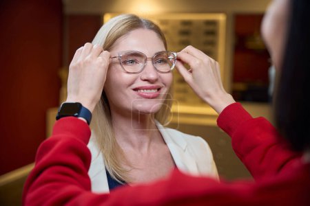 Consultant tries on glasses in a stylish frame for a client, a woman in a red pullover