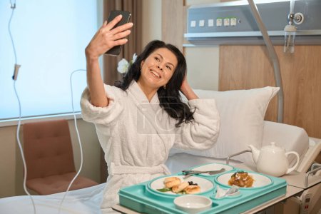 Lady takes selfie at breakfast in modern hospital ward, she uses mobile phone